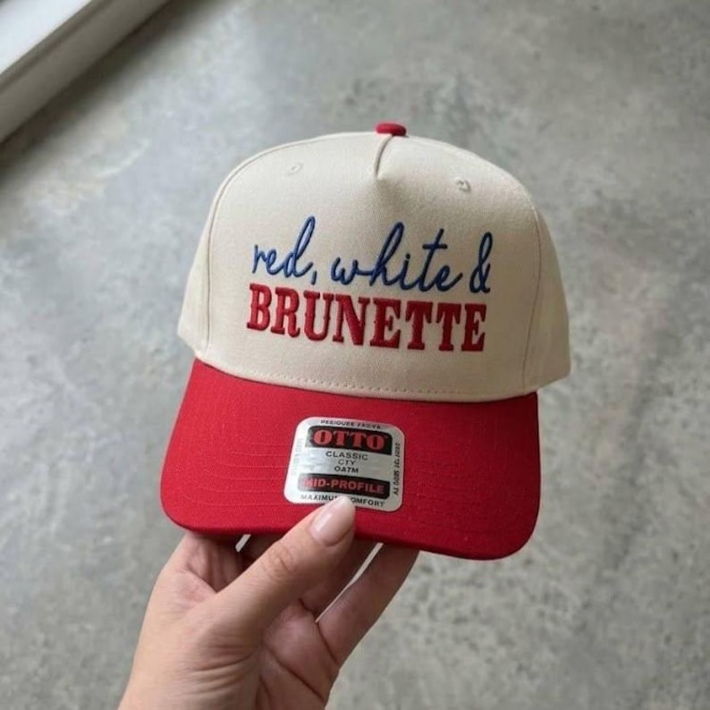 Peut inclure: Casquette de baseball avec une visi&egrave;re rouge et une couronne cr&egrave;me. Le texte "red, white & BRUNETTE" est brod&eacute; sur le devant en bleu et rouge. La casquette a un bouton rouge sur le dessus et une &eacute;tiquette sur la visi&egrave;re.