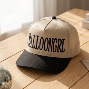 May include: A beige baseball cap with a black brim. The cap features the word "BALLOONGRl" embroidered in black. The cap is sitting on a wooden surface.