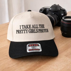 May include: A cream-colored baseball cap with a black brim. The text "I TAKE ALL THE PRETTY GIRLS PHOTOS" is embroidered in black. A camera and lens are in the background.
