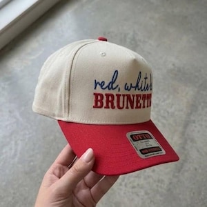 Peut inclure: Casquette de baseball beige et rouge avec le texte "red, white, BRUNETTE" brod&eacute; sur le devant. La casquette a une visi&egrave;re rouge et un bouton rouge sur le dessus. La casquette est tenue par une personne.