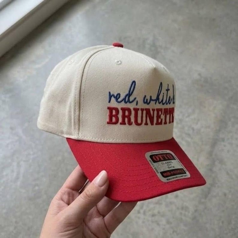 Peut inclure: Casquette de baseball beige et rouge avec le texte "red, white, BRUNETTE" brod&eacute; sur le devant. La casquette a une visi&egrave;re rouge et un bouton rouge sur le dessus. La casquette est tenue par une personne.