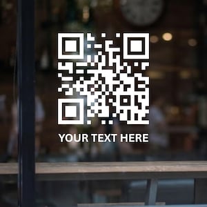 May include: A white QR code and the text "YOUR TEXT HERE" on a glass surface. The background is blurred, suggesting a cafe or shop interior with wooden features and a clock.