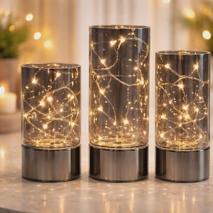 May include: Three decorative glass cylinders with warm white fairy lights inside. The cylinders have a silver base and are of varying heights. The lights create a soft, ambient glow, perfect for home decor or special events.