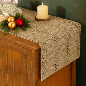 May include: A tan burlap table runner with a decorative open weave pattern. A lit candle sits on a wooden coaster. Christmas ornaments and greenery are arranged on the wooden cabinet. A teacup and saucer are also visible.
