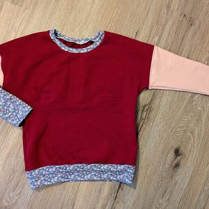 Könnte beinhalten: Ein weinrotes Sweatshirt mit pfirsichfarbenen Ärmeln und floral gemusterten Bündchen. Der Halsausschnitt und der Saum weisen ebenfalls das Blumenmuster auf. Das Kleidungsstück ist aus weichem Material gefertigt und für den Freizeitgebrauch geeignet.