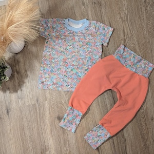 Könnte beinhalten: Ein zweiteiliges Baby-Outfit. Das Oberteil ist ein kurzärmeliges T-Shirt mit hellblauem Kragen und einem Blumenmuster in Blau-, Rosa- und Grüntönen. Die Hose ist korallenfarben mit floralen Bündchen, passend zum Muster des Shirts.