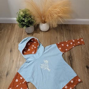 Könnte beinhalten: Hellblauer Kapuzenpullover mit rostfarbenen Ärmeln und floralen Akzenten. Die Innenseite der Kapuze ist mit weißen Blumen auf rostfarbenem Hintergrund gemustert. Auf der Vorderseite ist ein weißes Blumendesign gestickt.