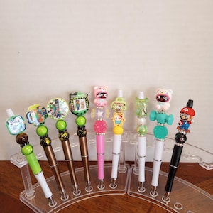 May include: A collection of decorative pens with various character toppers, including cartoon figures and designs. The pens are displayed in a clear plastic stand on a wooden surface. The pens have different colored barrels and toppers.