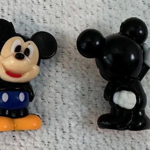 May include: Two small plastic Mickey Mouse figurines. The left figure shows a full-colour Mickey with a black head, ears, and body, blue shorts with white buttons, and yellow shoes. The right figure is a solid black silhouette of Mickey.