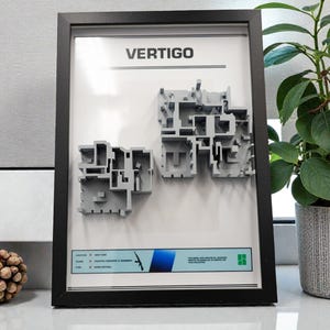 May include: Framed artwork featuring the word "VERTIGO" at the top. Below, two gray architectural models are displayed. A blue and white banner at the bottom includes text and a gun graphic. The frame is black.