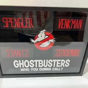 May include: A black framed picture with a Ghostbusters theme. It features the Ghostbusters logo, the iconic no ghost symbol, and the names of the four Ghostbusters: Spengler, Venkman, Stantz, and Zeddemore. The text "Who You Gonna Call?" is also included.