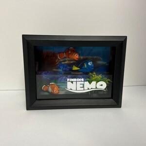 May include: A framed artwork featuring characters from the animated film "Finding Nemo." The scene includes Nemo, Dory, and other underwater elements. The title "Finding Nemo" is displayed prominently. The frame is black.