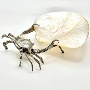 May include: A decorative crab-shaped sculpture made of silver-toned metal, paired with a large, iridescent white shell. The crab has detailed claws and legs, and the shell is supported by the crab's claws. The sculpture is on a white surface.
