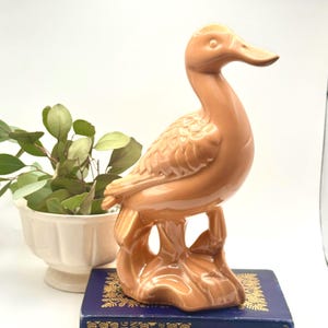 May include: A ceramic duck figurine in a warm peach color. The duck is perched on a stylized base, with detailed feather textures. The figurine is placed on a blue book, with a white planter and greenery in the background.