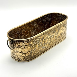 May include: A vintage brass planter with an oval shape and intricate floral detailing. The planter features a decorative pattern of flowers and leaves, with a small handle on one side. The brass has a warm, aged patina.