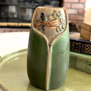 May include: A green and brown ceramic vase with a unique, stylized design. The vase features a central panel with a relief of a bird and branch. The vase has a smooth, glossy finish and is likely used for decorative purposes.