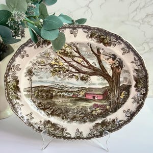 May include: An oval ceramic serving platter featuring a cream-colored background with a dark brown transferware border of grape leaves. The center depicts a landscape with a large tree, a red barn, rolling hills, and baskets of harvested fruit.