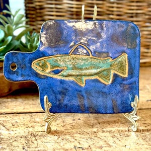 May include: A rectangular ceramic plaque with a handle and a circular hole, featuring a glossy deep blue glaze with a speckled texture. A raised, embossed relief of a fish in shades of green and gold is centered on the surface of the plaque.