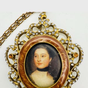 May include: A vintage-style pendant necklace featuring an oval portrait of a woman. The portrait is framed in a brown and gold setting, surrounded by clear and amber-colored crystals. The necklace has a gold-colored chain.