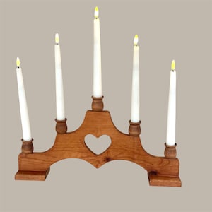 May include: A wooden candle holder with five white candles. The holder is made of light brown wood and features a heart-shaped cutout in the center. The candles are lit with yellow flames.