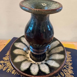 May include: A ceramic candle holder with a dark blue and brown glaze. The holder has a flared top and a round base with a floral design in white and gray. The candle holder is approximately 5 inches tall.