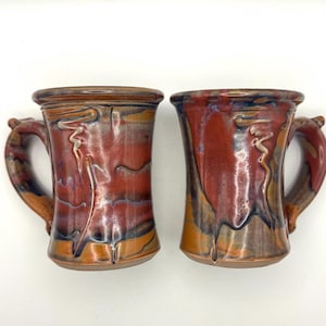 May include: Two handmade ceramic mugs with a rustic design. The mugs feature a rich blend of red, brown, and orange hues with a glossy finish. Each mug has a unique handle and a decorative swirl design on the front.
