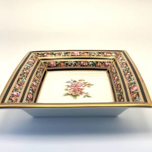 May include: Square porcelain dish with a floral design. The dish has a cream-colored interior with a central floral arrangement. The rim features a black and gold border with floral and leaf patterns. The dish is a decorative piece.