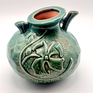May include: A small, round, teal-glazed ceramic vase featuring a textured, hammered surface and a raised, embossed floral design on the front. The vase has a wide, oval-shaped opening with a terracotta-colored interior and two small handles.