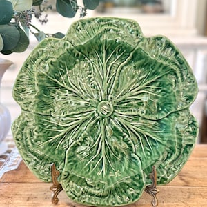 May include: A glossy green ceramic plate shaped like a cabbage leaf with intricate, raised vein details radiating from the center. The plate features a scalloped edge and is displayed upright on a small, decorative metal stand with ornate feet.