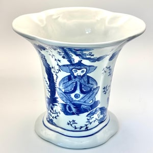 May include: A white and blue porcelain vase with a flared rim and a wide base. The vase features a blue illustration of a monkey wearing a hat and a vest, surrounded by floral and tree branch designs. The vase is approximately 8 inches tall.