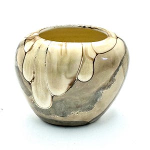 May include: A small, round ceramic vase with a wide opening. The exterior features a marbled design in shades of beige, brown, and gray, with a glossy finish. The interior is a solid yellow color.