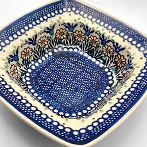 May include: A square ceramic bowl featuring a cream-colored base with a dense pattern of dark blue dots in the center. The rim is decorated with a repeating motif of brown and white flowers, green leaves, and blue accents on a cream background.