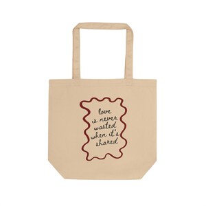 May include: Beige tote bag with a maroon, wavy-edged rectangular design. The design features the text "love is never wasted when it's shared" in a handwritten font. The bag has two beige handles.