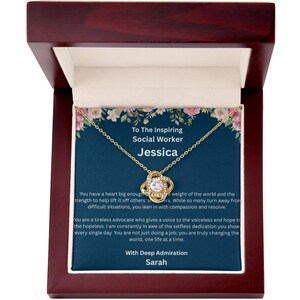 May include: A gold-colored necklace with a knot pendant, presented in a dark wooden box. The pendant features a central clear stone surrounded by smaller stones. The box contains a card with text honoring a social worker named Jessica.