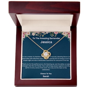 May include: A gold-colored necklace with a pendant in a dark wooden jewelry box. The pendant features a clear stone surrounded by a knot design. The box contains a card with text that reads "To The Amazing Bartender Jessica".