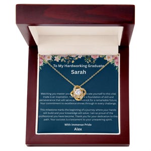 May include: A gold-colored necklace with a pendant in a wooden jewelry box. The pendant features a knot design with a central clear stone. The box contains a card with text that reads "To My Hardworking Graduate Sarah."