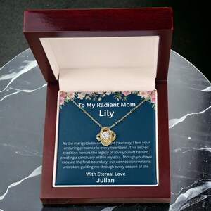 May include: A gold-tone pendant necklace with a central cubic zirconia stone in a knot setting, presented in a dark wood box. The display card features floral borders, the text To My Radiant Mom Lily, and a message ending with With Eternal Love Julian.