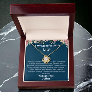 May include: A gold-tone necklace with a central knot pendant featuring a clear stone, presented in a dark wood box. The display card inside features a floral border, a dark blue background, and text addressed to Lily from Julian with a message.