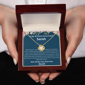 May include: A gold-tone necklace with a central knot pendant featuring a clear stone, presented in a dark wood box. The interior card displays floral accents and text: To My Brilliant Accountant Sarah, followed by a message and With All My Love And Pride David.