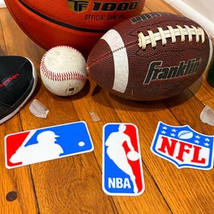 Può includere: Una collezione di attrezzature sportive tra cui un pallone da basket rosso e nero, un pallone da football americano marrone e una palla da baseball bianca. Sono presenti anche adesivi con i loghi MLB, NBA e NFL.