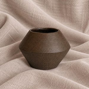 May include: A dark brown ceramic vessel with a wide, angular midsection that tapers toward a narrow base and a circular opening at the top. The surface of the object features a matte, textured finish with subtle speckles throughout the dark clay.