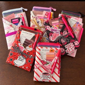 May include: A collection of five fabric pouches featuring various patterns including hearts, stripes, and floral prints. Each pouch contains a reading list card, colorful sticky note tabs, and a pen, decorated with stickers and tied with twine.