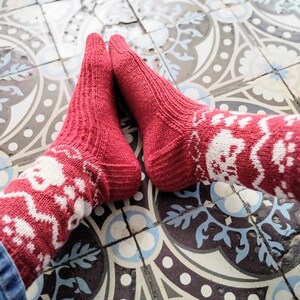 May include: A pair of red knitted socks with a white pattern of skulls and paw prints. The socks are worn on a patterned tile floor. The socks are made of a soft, warm material.