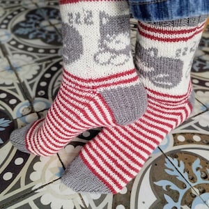 Könnte beinhalten: Gestrickte Socken in Grau, Rot und Weiß. Der Fußbereich hat rot-weiße Streifen, mit einer grauen Ferse und Spitze. Der obere Teil hat einen weißen Hintergrund mit einem grauen Katzenmuster und roten Streifen, für ein gemütliches Aussehen.