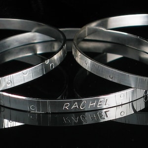 May include: Three silver bangle bracelets with engraved names and measurements. The bracelets are stacked on top of each other. The names engraved on the bracelets are "Jameson", "Rachel", and "One must not speak of the dark mark".