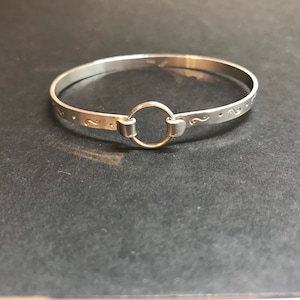 May include: A silver bangle bracelet with a circular clasp. The bracelet is adorned with small, decorative swirl patterns and dots. The clasp is a simple, round design. The bracelet is made of a shiny, metallic material.
