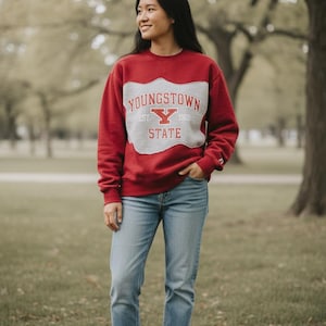 May include: A red sweatshirt with a gray panel featuring the text "YOUNGSTOWN STATE" and a "Y" logo. The person is wearing light blue jeans and white sneakers. The background is a park with trees.