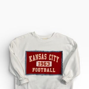 May include: White sweatshirt with a red patch featuring "KANSAS CITY" above "1963 FOOTBALL" in white lettering. The patch is outlined with a blue denim-like material. The sweatshirt has long sleeves and a crew neck.