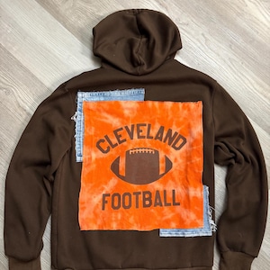 May include: Brown hooded sweatshirt with a tie-dye orange and brown patch on the back. The patch features the words "Cleveland Football" and a football graphic. Patches of denim are sewn on the back.