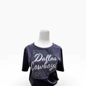 May include: A navy blue t-shirt featuring the text "Dallas Cowboys" in white script, overlaid on a gray outline of the state of Texas. A white star is below the text. The shirt has short sleeves.
